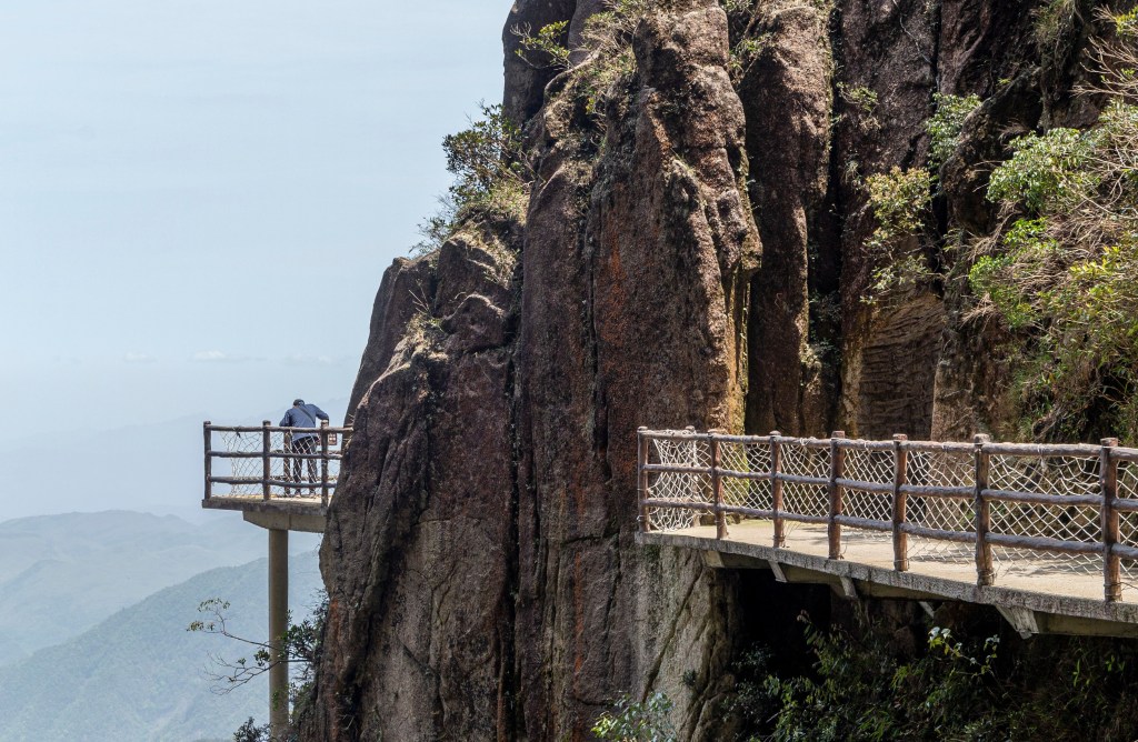 A person standing on a wooden walkway extending from a rocky cliff, overlooking a mountainous landscape in the background.